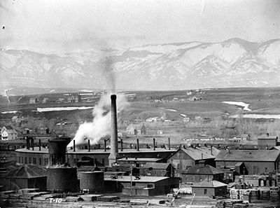 View of Burlington railyard in Sheridan, showing plume of steam from roundhouse and stacks of raw and treated railroad ties adjacent to the tie-treatment plant. Courtesy of Wyoming Room, Sheridan County Fulmer Public Library.
