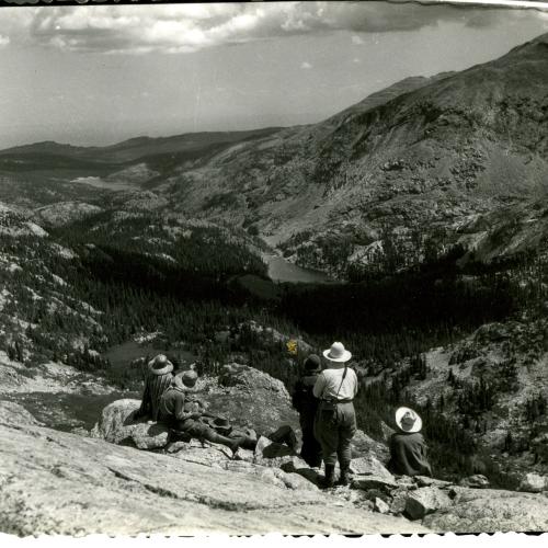 A group of four or five people above timberline look down an alpine valley at a lake in the distance