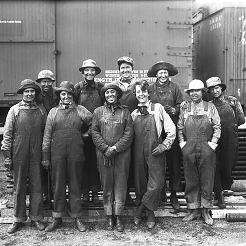 A group of ten women in overalls pose in front of railroad cars