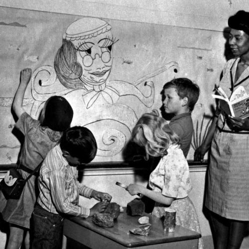 Liz Byrd stands with four elementary school studnets looking at a wall chart