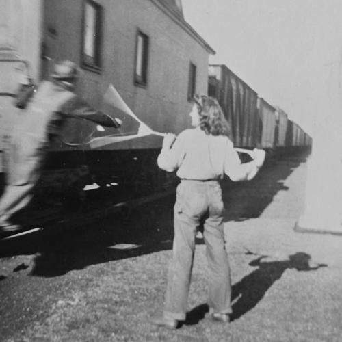 a railroad worker leans from the caboose to grasp at a long rake-like stick being held by a woman standing next to the track