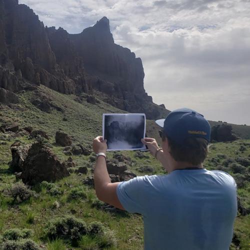 Man holds up a photocopy of an old photograph in front of the Organ Pipes landscape