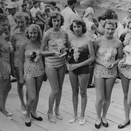 Seven women in one-piece bathing suits pose, standing in a row