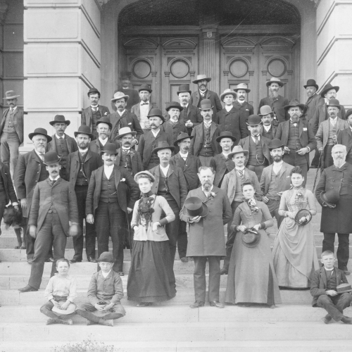 Delegates to the Wyoming Constitutional convention gather on the steps of the Wyoming Capitol building. In front there are three women who served as secretarial staff during the convention. There are also several children and a dog posing alongside the delegates.