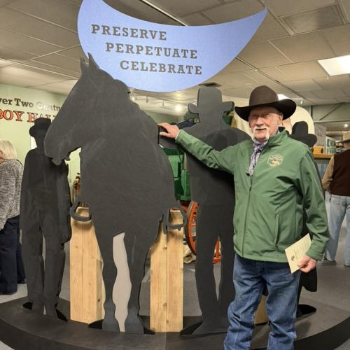 A man in a cowboy hat has his hand on the cut out of a horse, under the "Preserve Perpetuate Celebrate" sign