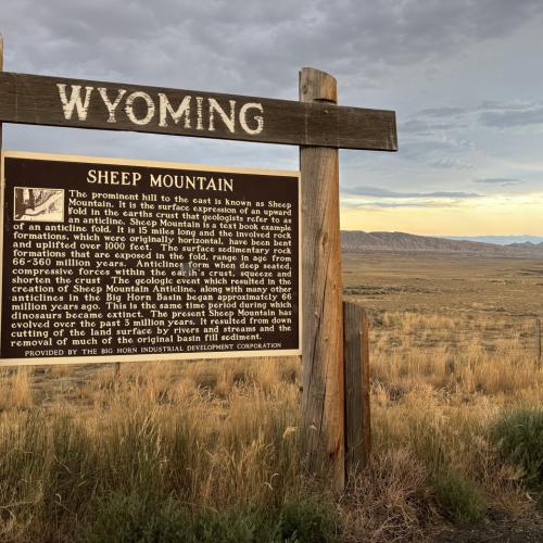 roadside sign hung from a wood crossbar that says, "Wyoming;" text on sign is titled "Sheep Mountain" and describes the geology of the areay