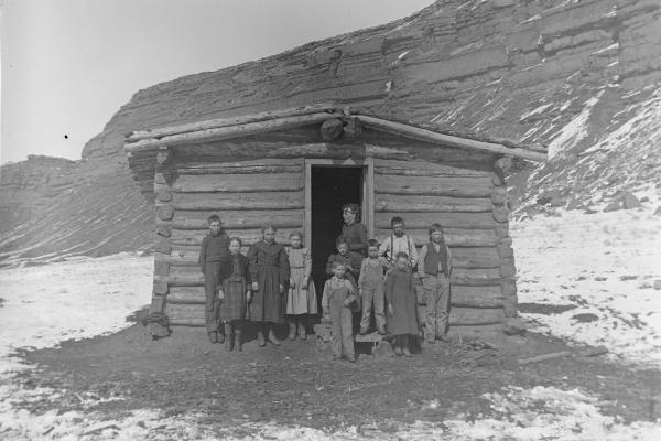 black and white photo of a one-room log building with ten children and a women standing out front. 