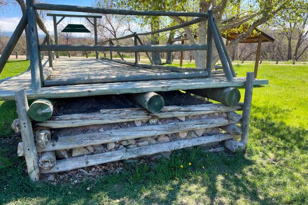 Stacked logs and rocks underpinning a wooden ramp with railings, set on a lawn