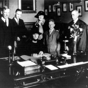 Nellie Tayloe Ross, dressed in mourning black for her late husband, signs the Oath of Office as Wyoming governor on January 5, 1925, joined by family: Judge Saul Tayloe, William Bradford Ross, Mrs. George Tayloe, George Ross, and Chief Justice Charles N. Potter. Courtesy Wyoming State Archives.