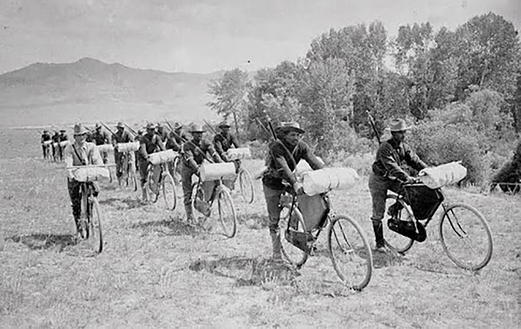 more than a dozen soldiers with rifles on their backs riding bicycles across the plains, each with large parcels of gear on the handlebars