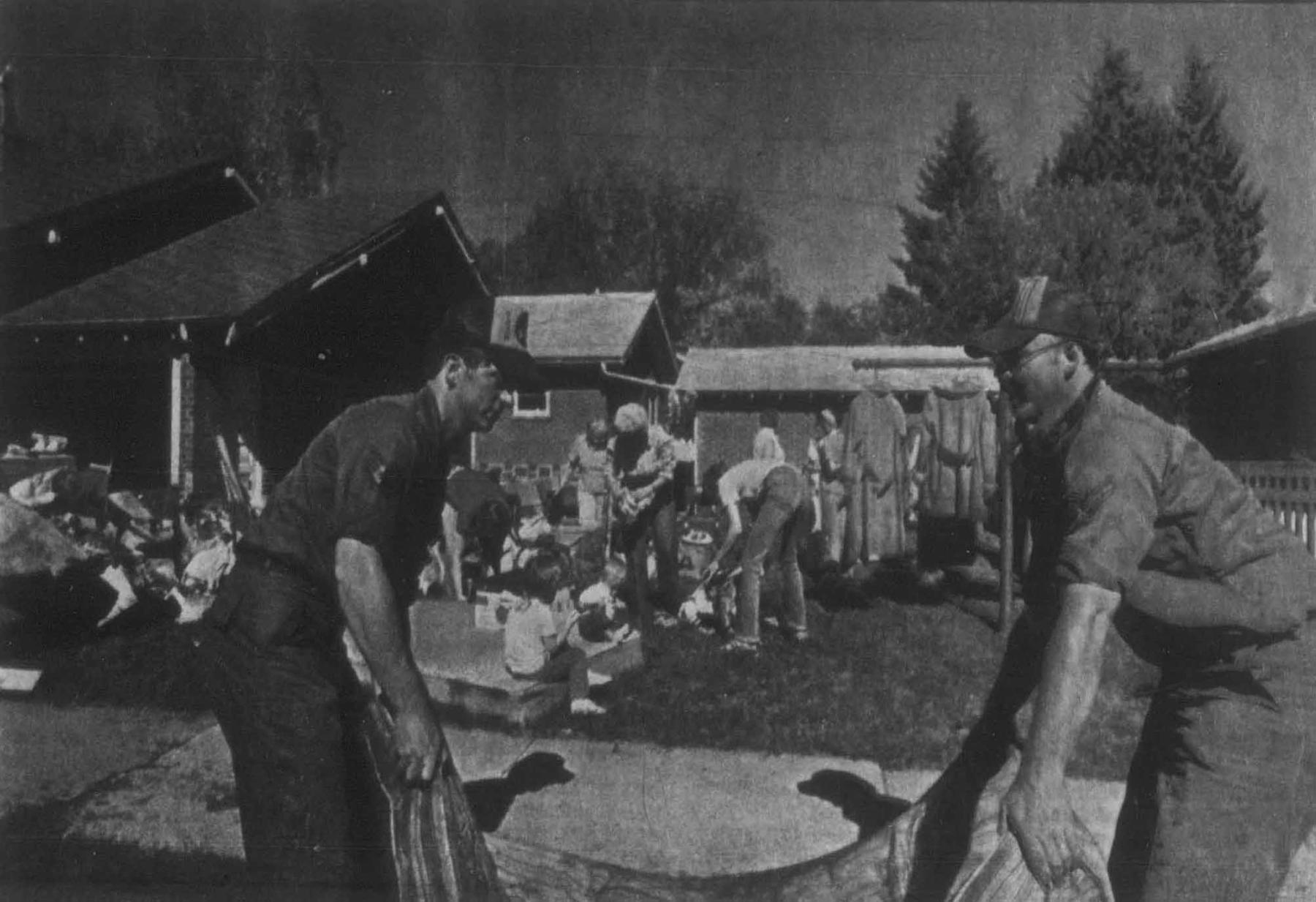 Air Force personnel SSgt Jeff Harrell (left) and AIC Gary Pontin help pump water and salvage belongings from a flooded basement. Two men in the foreground hold a tarp while a group of other peole are gathered in the bckground sorting through household articles outdoors