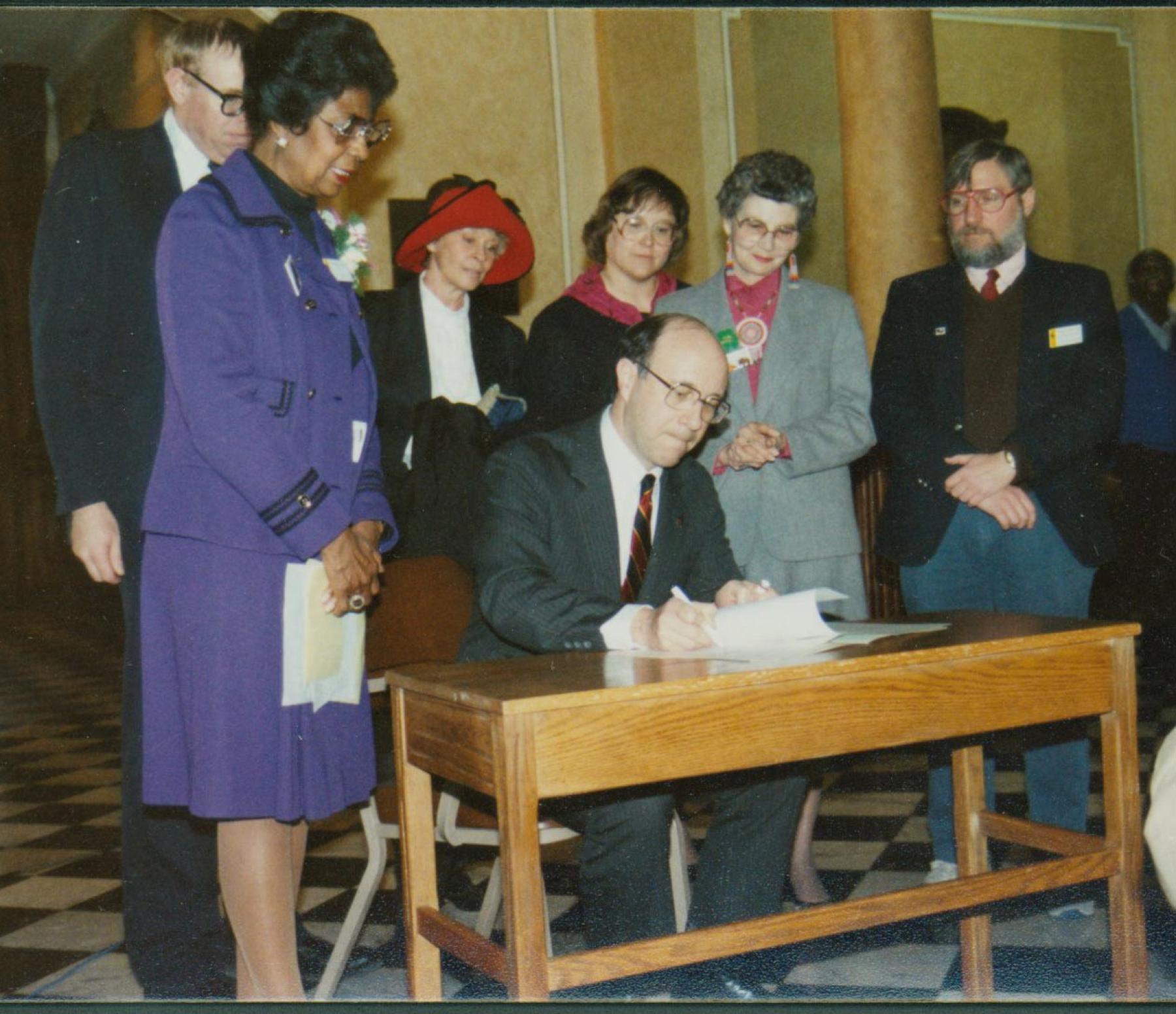 A group of women and men stand around a desk where a man is signing a document