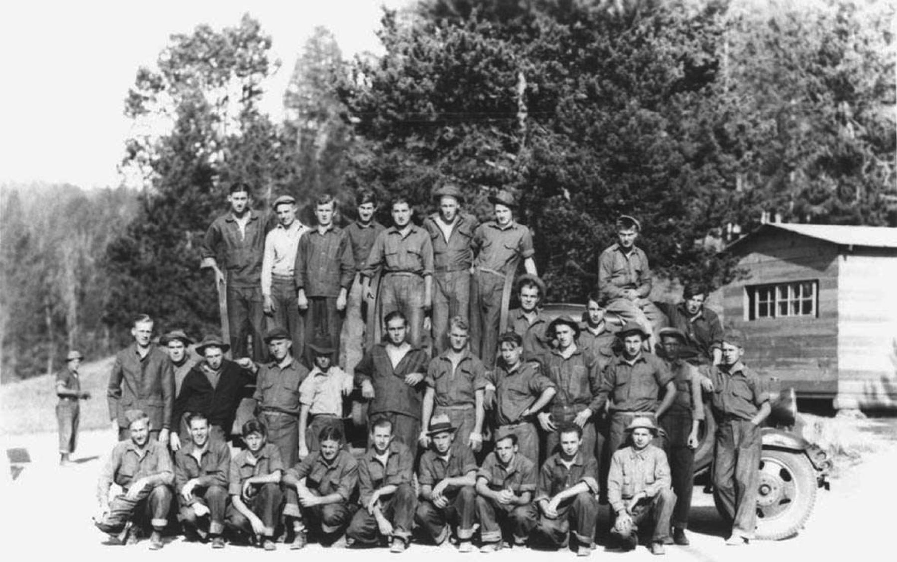 Three rows of young men in work clothing, some perched on a truck, pose near a cabin