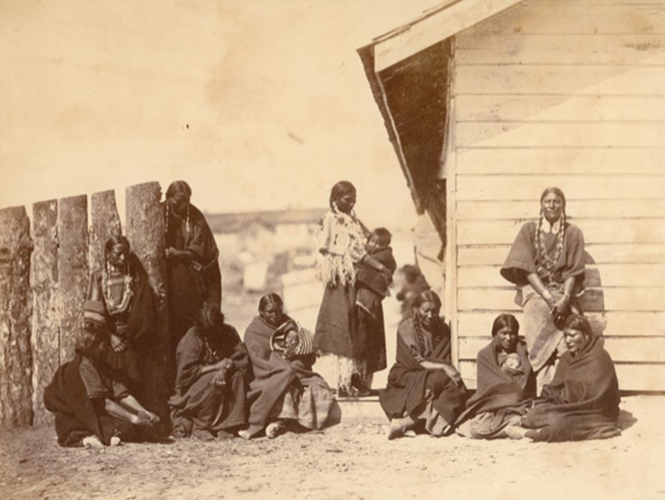 Lakota Sioux women, children and, against the right-hand end of the fence, a man, possibly the Oglala Lakota, High Wolf, at the Indian agent's quarters at Fort Laramie, 1868. Sherman collection of Gardner photographs; NAMI.