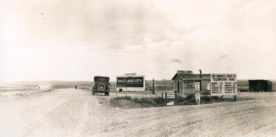 A small building stands at a gravel road intersection in a wide open plain. Sign atop the building says "Trail Inn/Free Campground." Road signs give distances: Green River, 30mi; Granger 2mi; Large sign says, "The Shortest Route to Yellowstone National Park" with distances Opal, 30mi; Kemmerer 46mi; Lava Hot Springs 182mi; Pocatello 220mi. A large billboard points to Salt Lake City and Bryce and Grand Canyons.