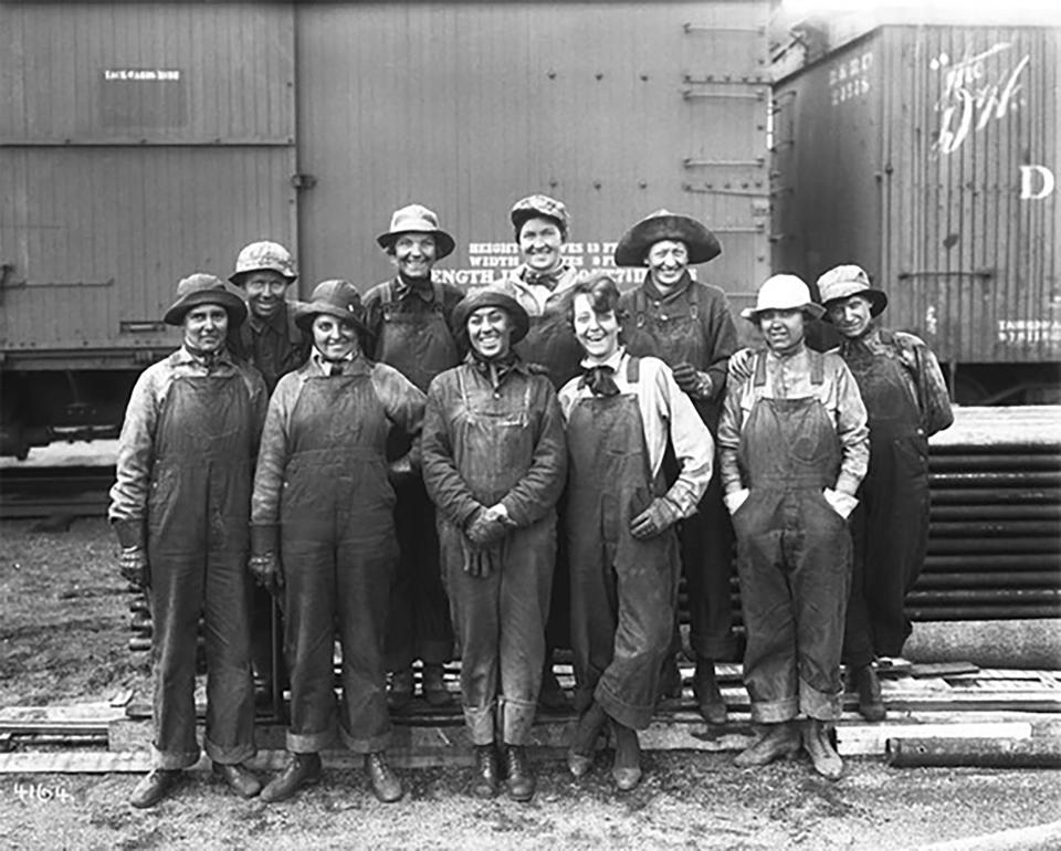 A group of ten women in overalls pose in front of railroad cars