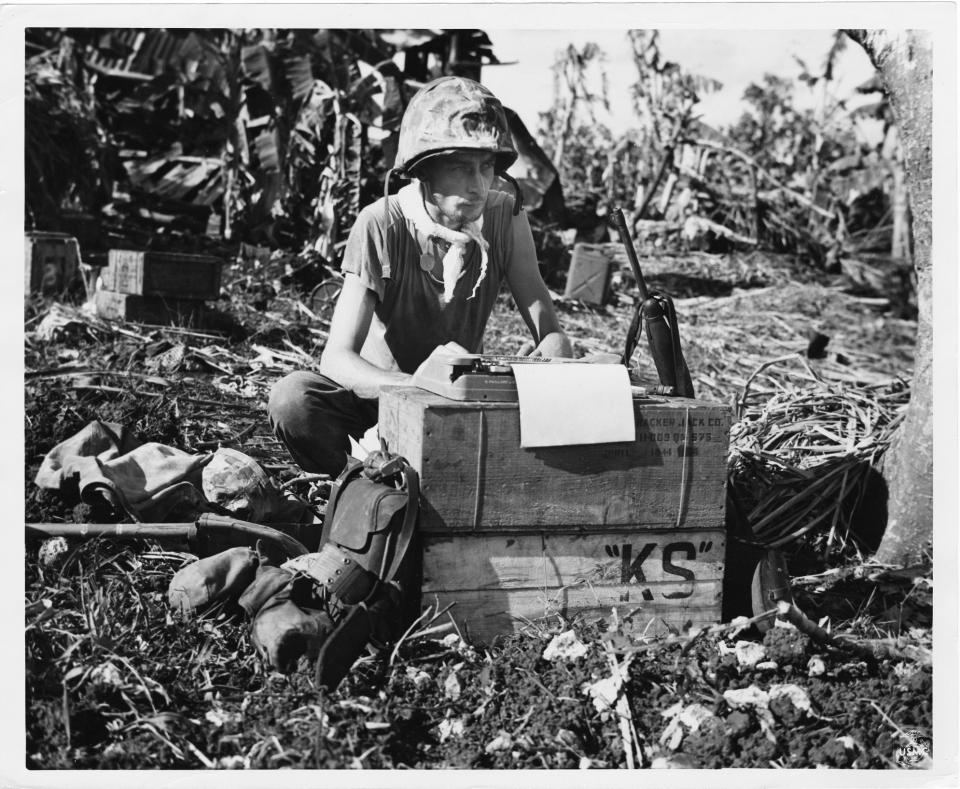 Man in combat fatigues and helmet sits in fromt of several boxes that prop up his small typewriter, outdoors in a place with tropical foliage.