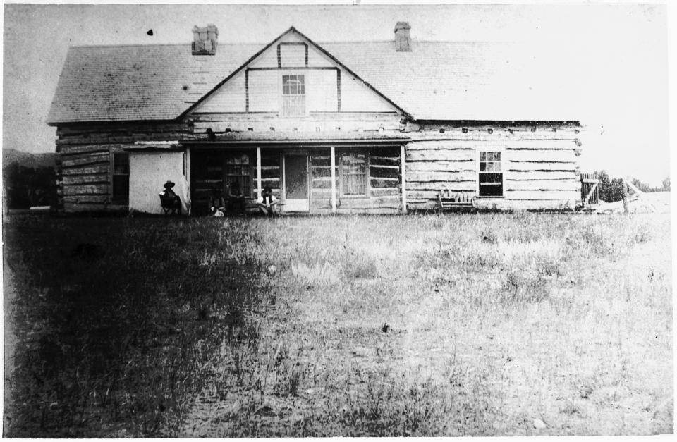 Large log house with a few people sitting on the front porch and in a chair in the front yard.