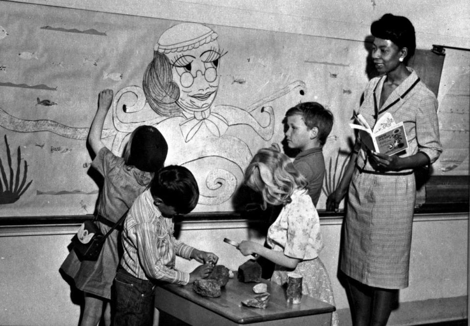 Liz Byrd stands with four elementary school studnets looking at a wall chart