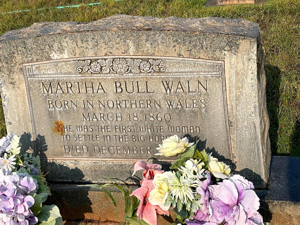 Headstone with the words, "Martha Bull Waln; Born in Northern Wales; March 18, 1860; She was the first white woman to settle in the Big Horn Basin; Died December [content obscured by artificial flower bouquet]"