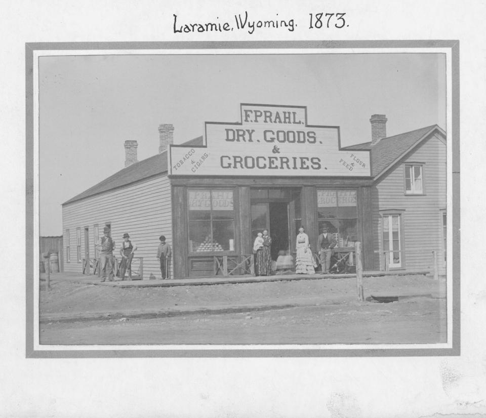 Photo, labeled "Laramie Wyoming 1873," shows wooden storefront with sign, "F. Prahl Dry Goods and Groceries / Tobacco and Cigars / Four and Feed." Several people stand on the wooden boardwalk in front.