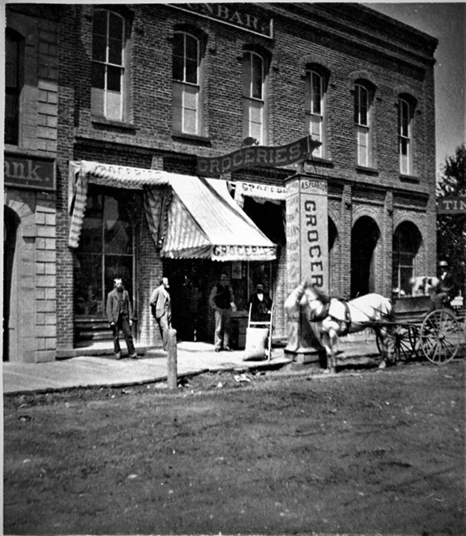 Storefront in a brick building along a boardwalk lining a dirt road. Ther are at leasr four signs adorning the entrance, all advertising "GROCERIES."