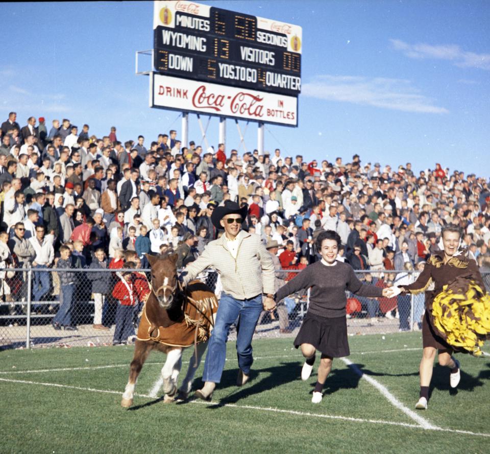 A young man in a cowboy hat, holding hands with two cheerleaders, leads a Shetland pony along the sidelines of a football field, with a scoreboad looming over the crowd in the bleachers 