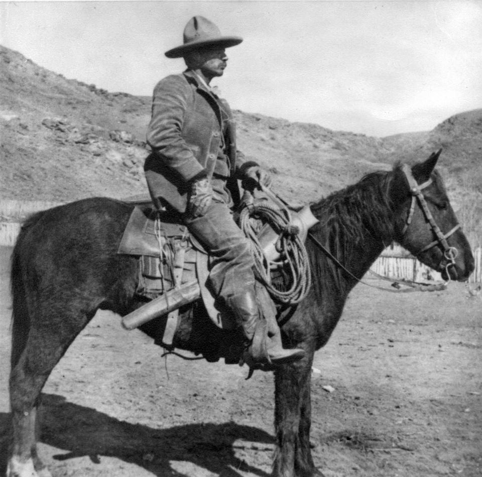 a man wearing a cowboy has, chaps, boots, and gloves, on a saddled horse, in profile