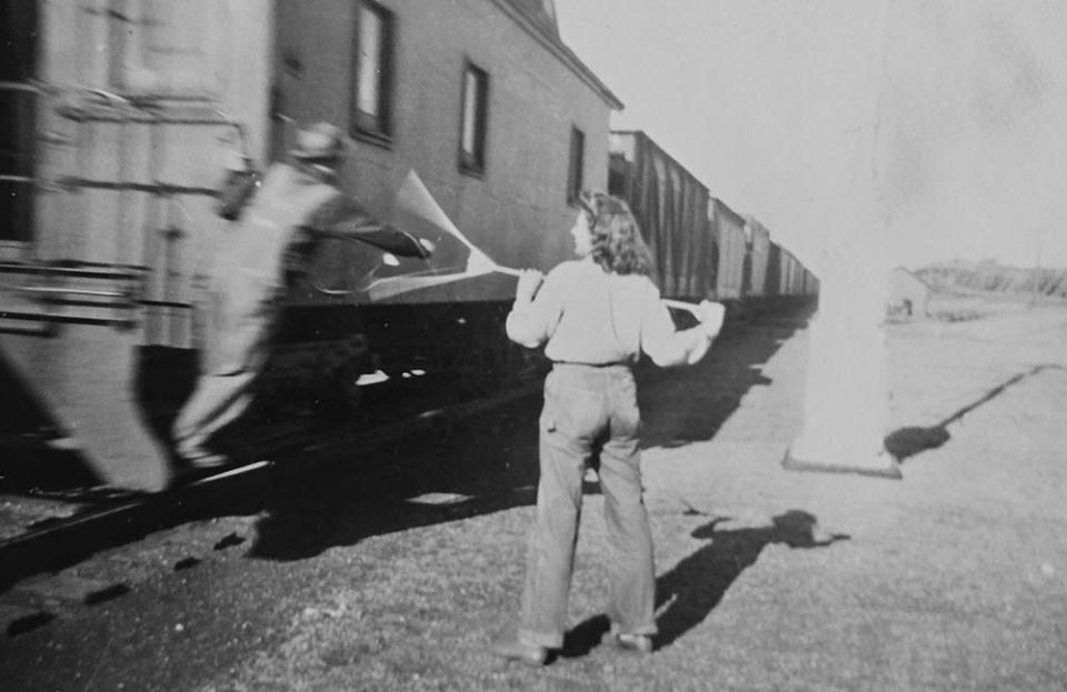 a railroad worker leans from the caboose to grasp at a long rake-like stick being held by a woman standing next to the track
