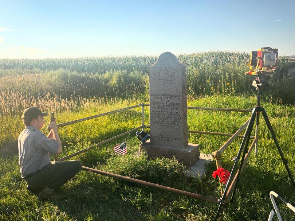 a man holding a piece of equipment squats at the edge of the railing around a stone monument. His large format camera is on a tripod nearby.