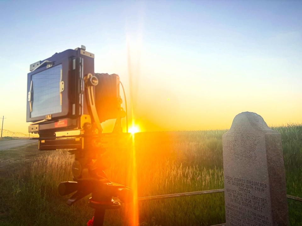 side view of a large-format camera near a stone monument. Rays from the rising sun bean directly into the camera and partially obscure the camera and the monument