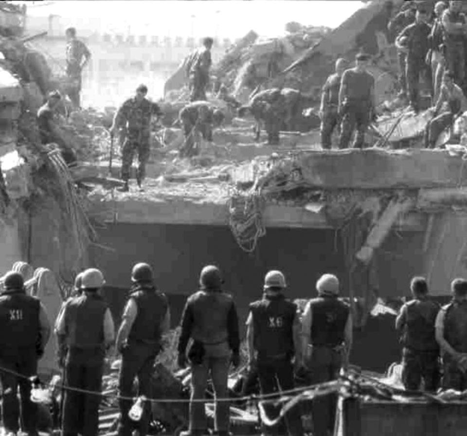 A line of helmeted men in vests observe the wreckage of a building where many other men bend over the rubble and look in various directions.