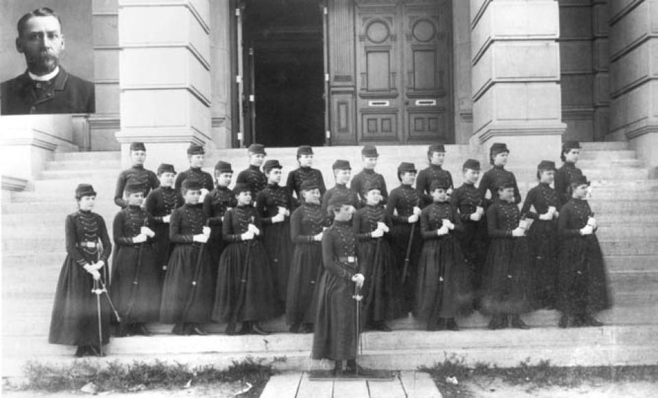 three lines of young women dressed in dark skirted military uniforms and caps, holding with swords or rifles, looking left while posed on the steps leading into a large building. An inswet at the upper left is a portrait of a man with a moustache