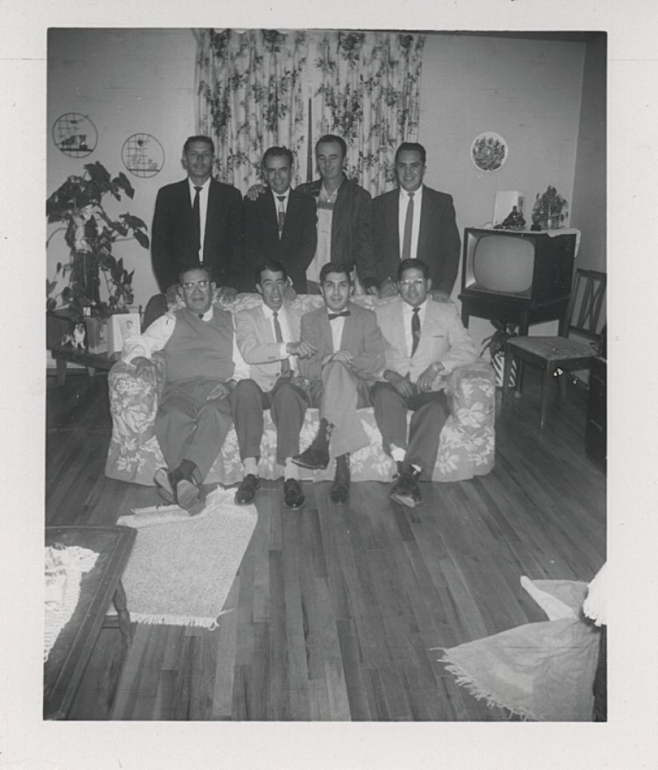 Eight men in coats and ties, four sitting on a sofa and four standing behind them, pose in a residence