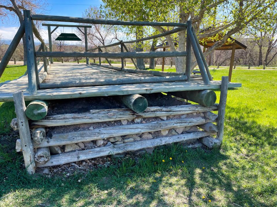Stacked logs and rocks underpinning a wooden ramp with railings, set on a lawn