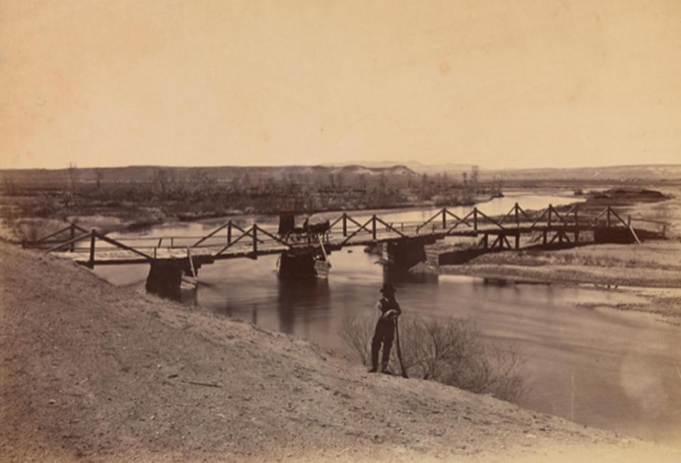 Wooden bridge spanning a river, with a man dressed in a dark hat, vest, and dark pants leans on a rifle in the foreground