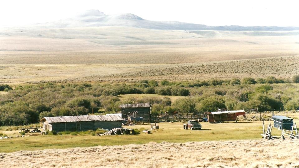 view of low-lying ranch buildings on a plain above a riverbed with rounded mountains in the distance