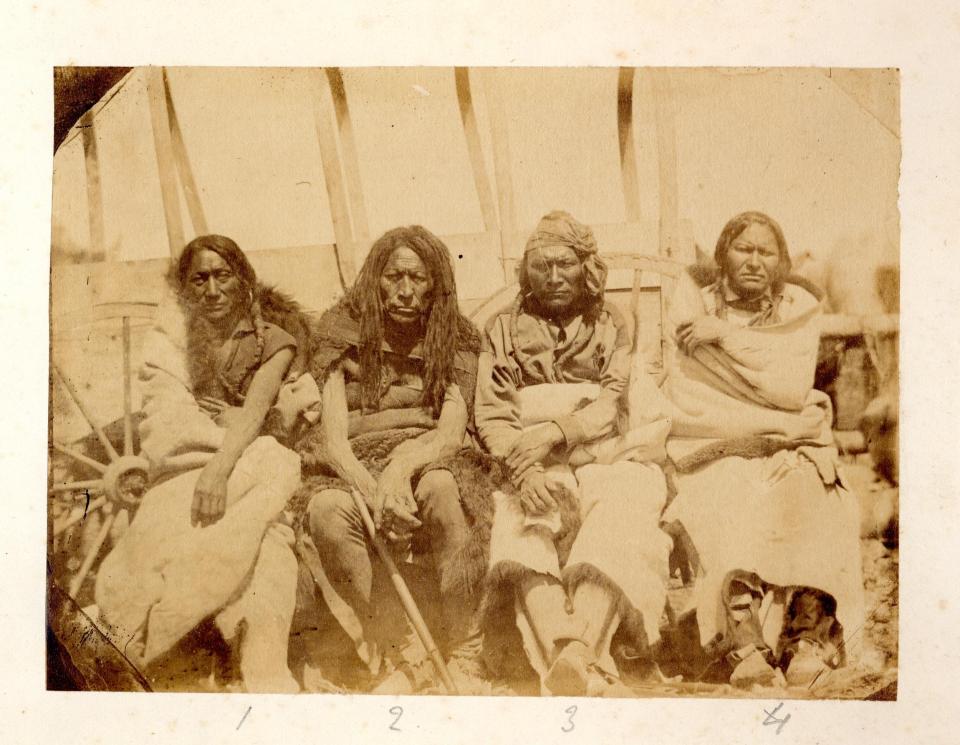 Four men seated in front of a covered wagon; three with dark hair in braids, the third with what appears to be dreadlocks. They are serious-faced, wearing robes and buckskin boots.