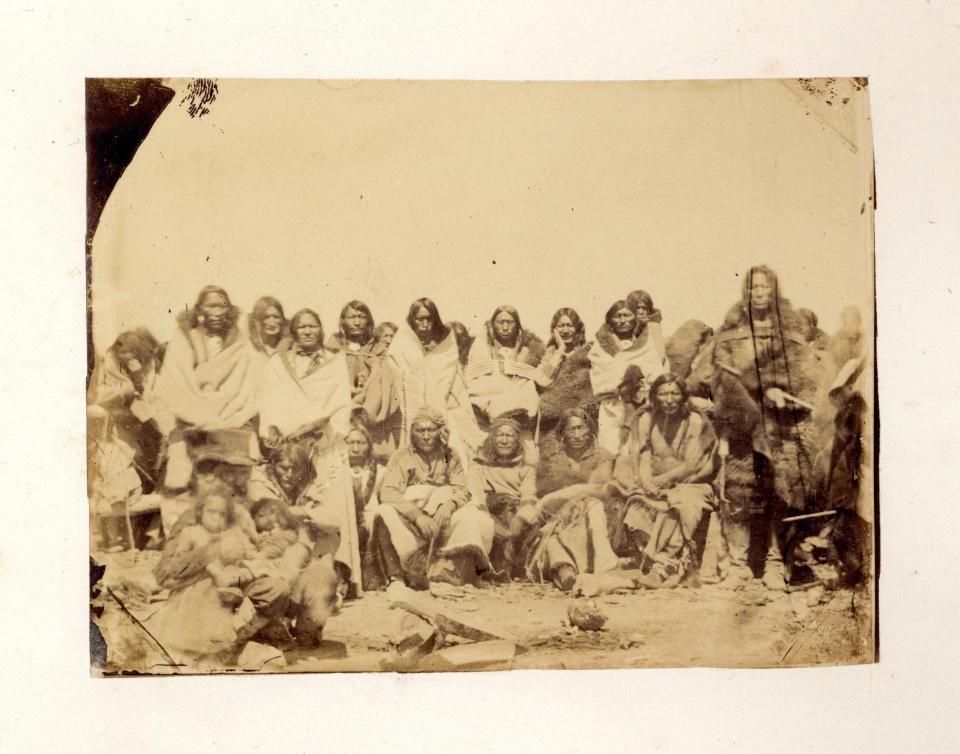 Group of about 15 Arapaho men an women, posed in a seated row in front, with others standing behind. They are unsmiling and most are dresssed in robes