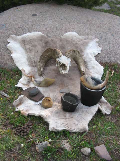 Mountain Shoshone culinary utensils and food, including sheephorn ladle, soapstone bowls, a horn-hafted stone knife, mano and metate and, in the horn bowl, rose hips--and wild mushrooms in the grass. The tools are replicas. Tory Taylor.