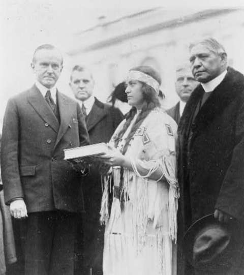 Cherokee activist Ruth Muskrat Bronson of the reformist Committee of One Hundred presents a book to President Calvin Coolidge while Sherman Coolidge looks away, 1923. Library of Congress.