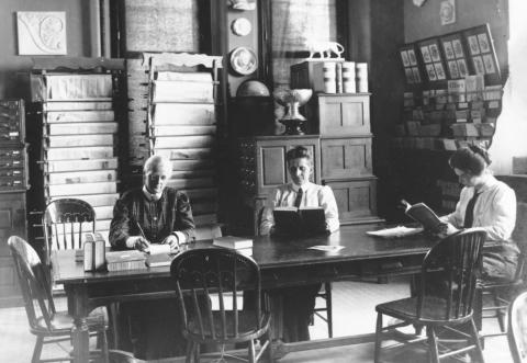 Three women in long dresses reading and writing, seated at a table in a room with cupboards and racks of papers