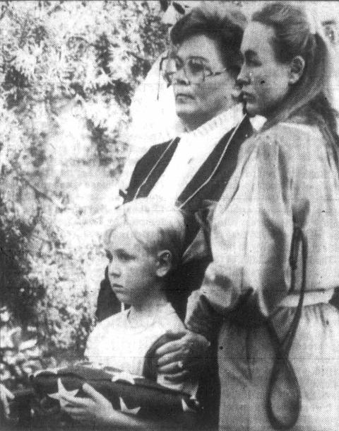 Eight-year-old Robert Van Alyne Jr. clutches the flag from his father’s casket, supported by his grandmother Barbara “Dolfe” Van Alyne. Two women stand behind a young boy who is holding a folded flag.