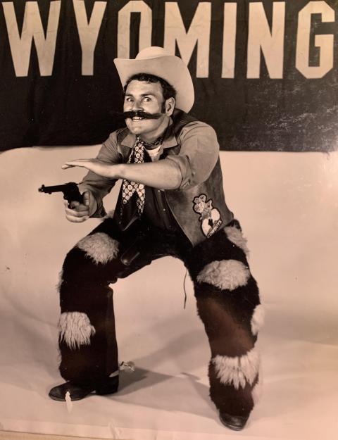 man in Pistol Pete costume poses crouching and aiming his pistol im front o a large "WYOMING" sign