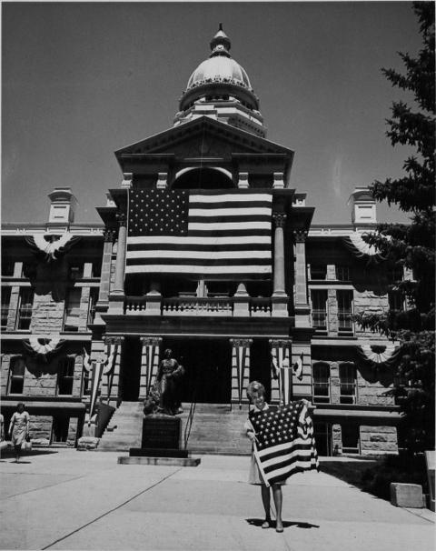 woman poses in front of the Wyoming State Capitol, holding and American flag. There is a much larger American flag displayed across the front of the building 