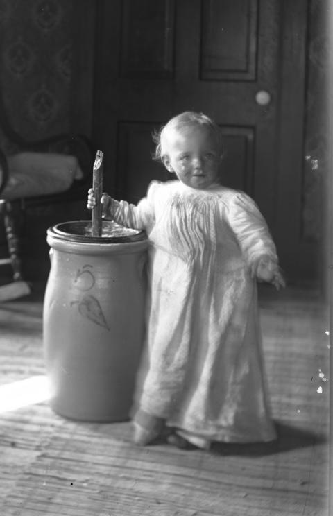 a child wearing a long white dress stands smiling holding the plunger of a butter churn