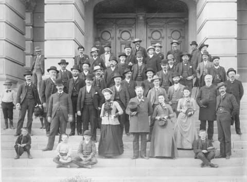 Delegates to the Wyoming Constitutional convention gather on the steps of the Wyoming Capitol building. In front there are three women who served as secretarial staff during the convention. There are also several children and a dog posing alongside the delegates.
