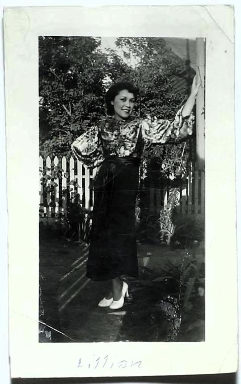 A dark haired woman in a dress with a floral blouse and high heels poses with one hand on her hip and the other on a wall