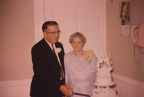 An elderly couple hold hands grasping a knife ready to slice a tiered wedding cake