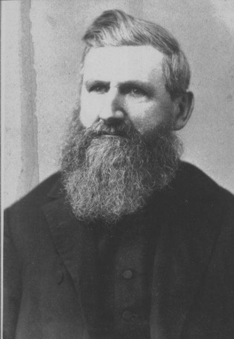 Head shot of a man in black with a dense ten-inch beard and moustache